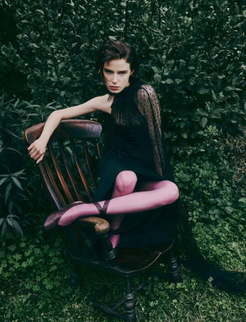 Woman in black dress and pink ballet shoes sits on wooden chair in lush green garden setting; fashion editorial photo.