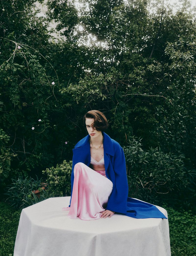 A woman in a blue coat sits on a table in a garden with lush green foliage. The woman is wearing a pink and white skirt.