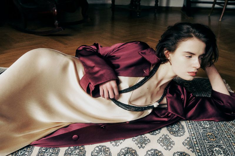 Brunette woman in a burgundy jacket and light yellow dress poses while lying on an ornate rug in a classically decorated room.
