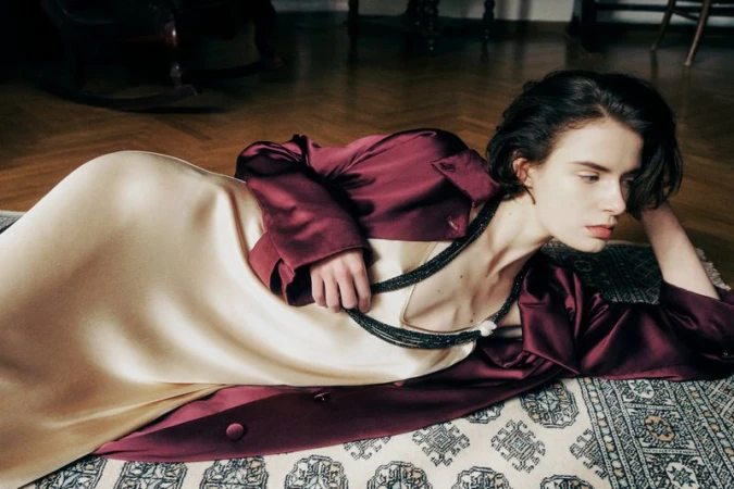 Brunette woman in a burgundy jacket and light yellow dress poses while lying on an ornate rug in a classically decorated room.