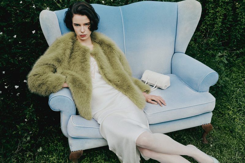 Elegant woman in a fuzzy olive coat poses in a blue vintage chair outdoors, holding a small purse, with lush green foliage.