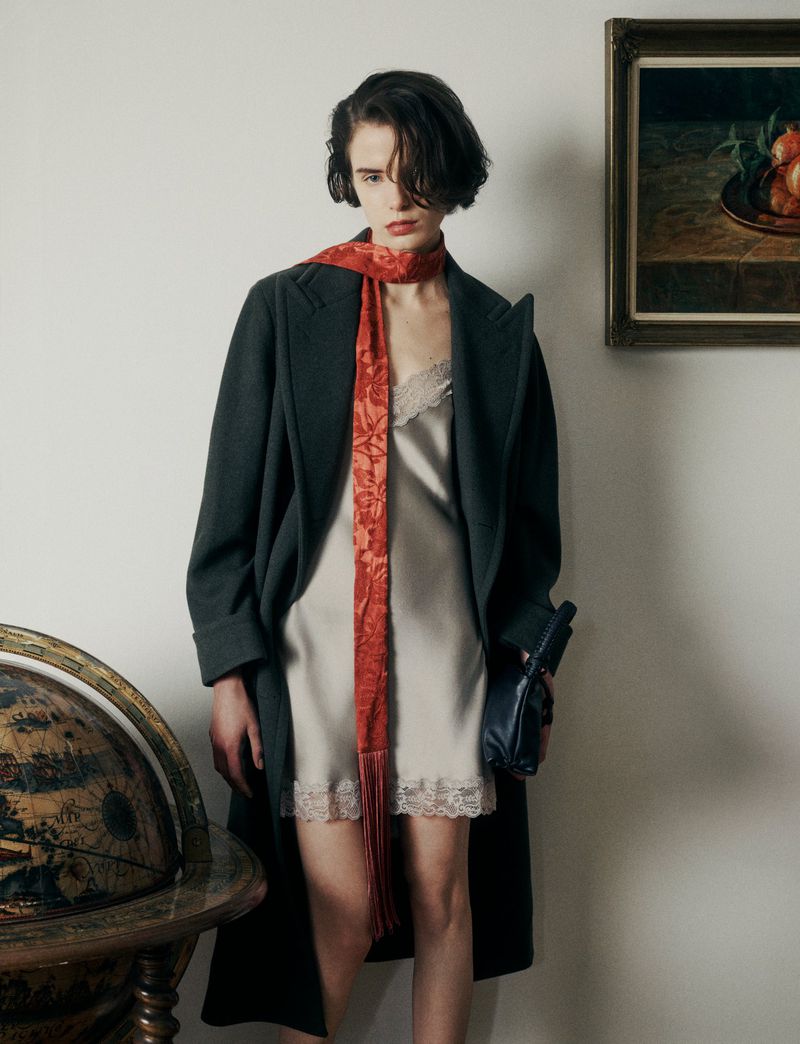 Woman in stylish coat and slip dress with a patterned scarf, posing indoors with a globe and framed artwork in the background.