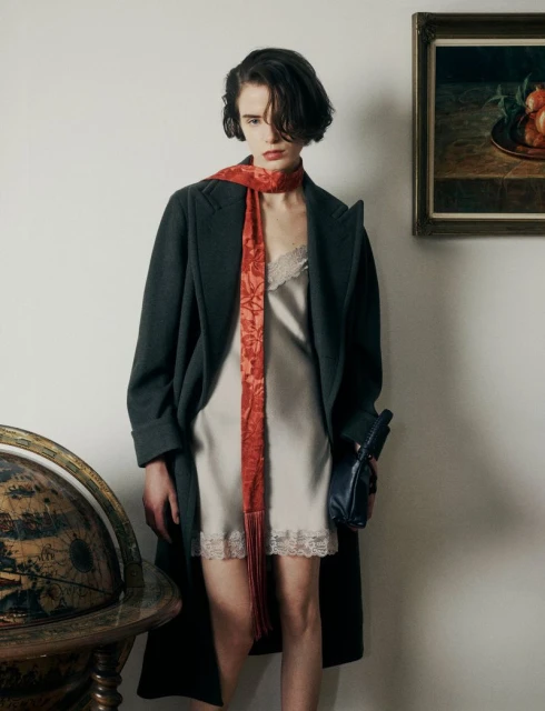 Woman in stylish coat and slip dress with a patterned scarf, posing indoors with a globe and framed artwork in the background.