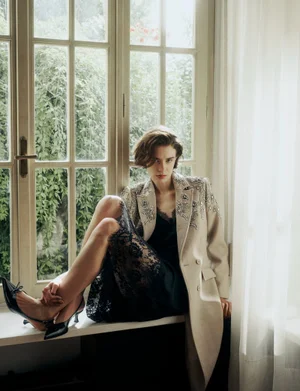 Model Greta Carnbring sitting on a window sill wearing an embellished beige coat and black lace slip dress for Vogue Greece.