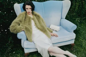 Greta Carnbring wearing an olive green fuzzy coat and white slip dress, seated in a light blue armchair on grass.