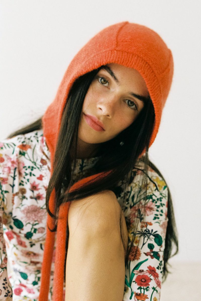 Portrait of a young woman with long brown hair, wearing a floral top and an orange knit hat with long ties against a white background.