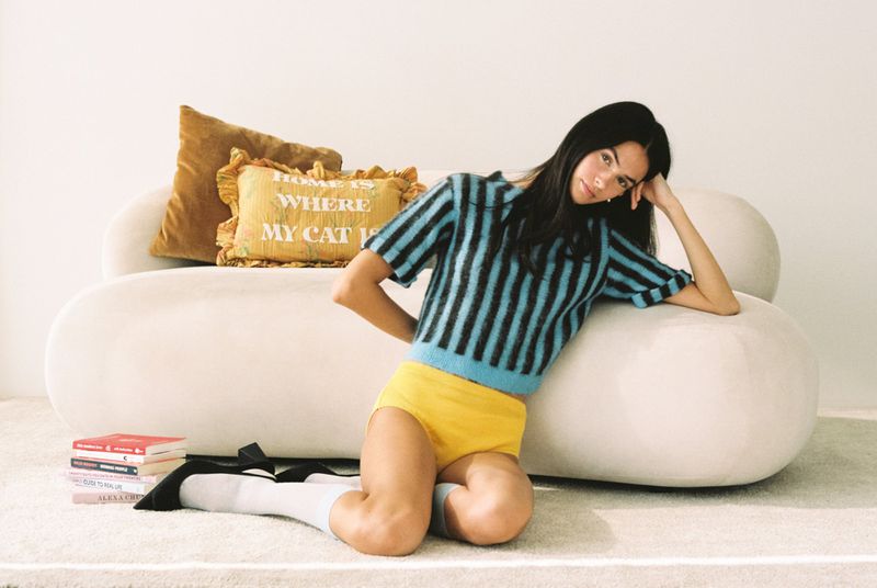 A young woman with long black hair, wearing a striped sweater and yellow shorts, poses on a white couch with decorative pillows indoors.