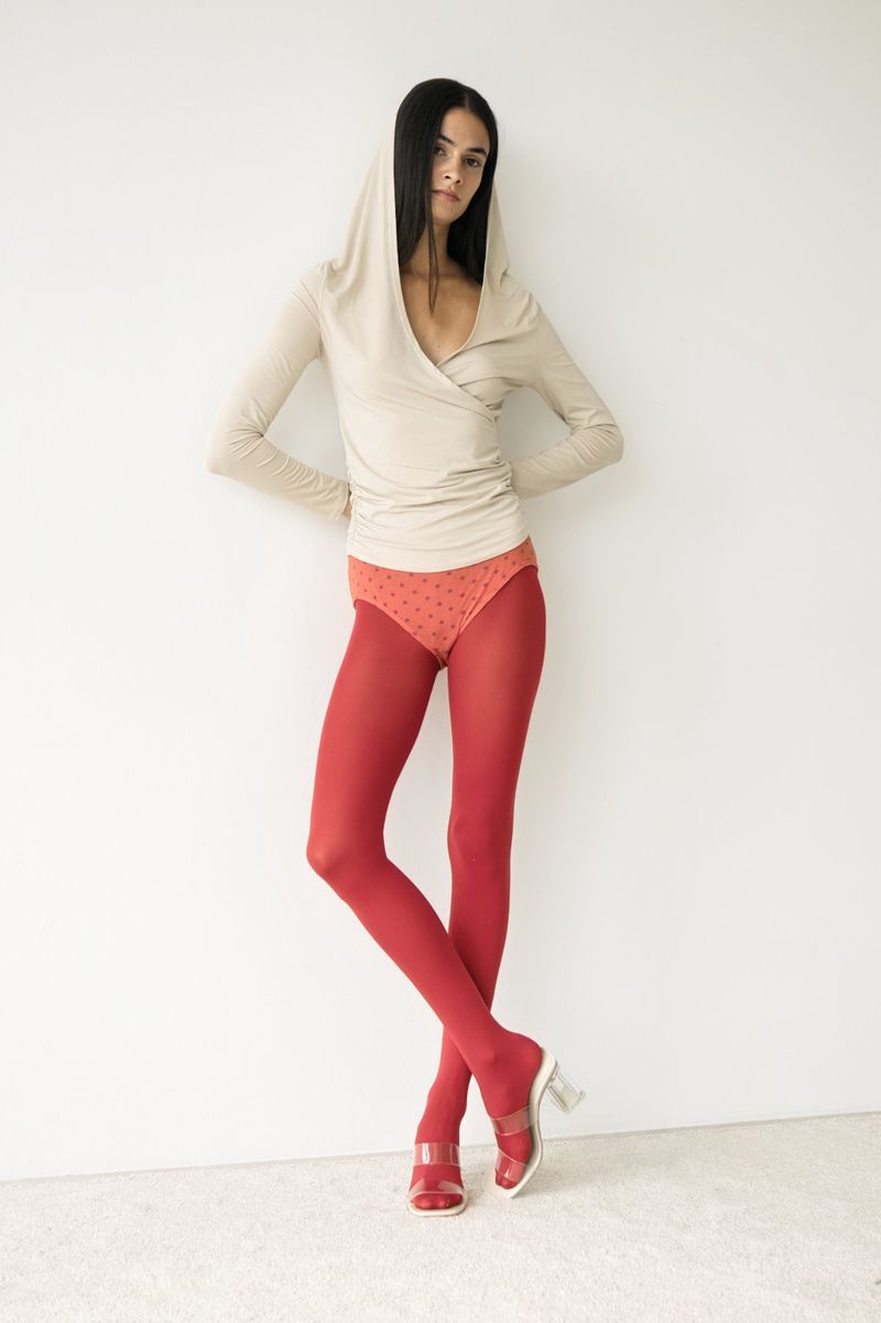 Fashion model poses in a beige hooded wrap top, red tights, and open-toed block heels against a stark white backdrop in a studio shot.