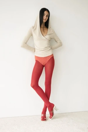 Fashion model poses in a beige hooded wrap top, red tights, and open-toed block heels against a stark white backdrop in a studio shot.