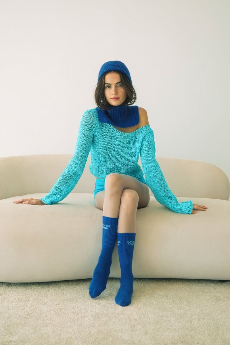 Young woman in a turquoise knit outfit with a matching beanie and blue socks, posing on a beige couch against a white wall backdrop.