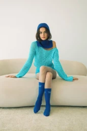 Woman in a bright turquoise open-knit sweater and blue beanie sitting on a curved cream sofa against a white background.