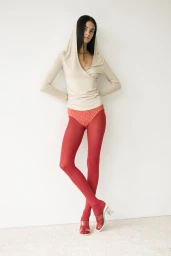 Model wearing a hooded beige wrap top and bold red tights posing full-length against a plain white studio wall.