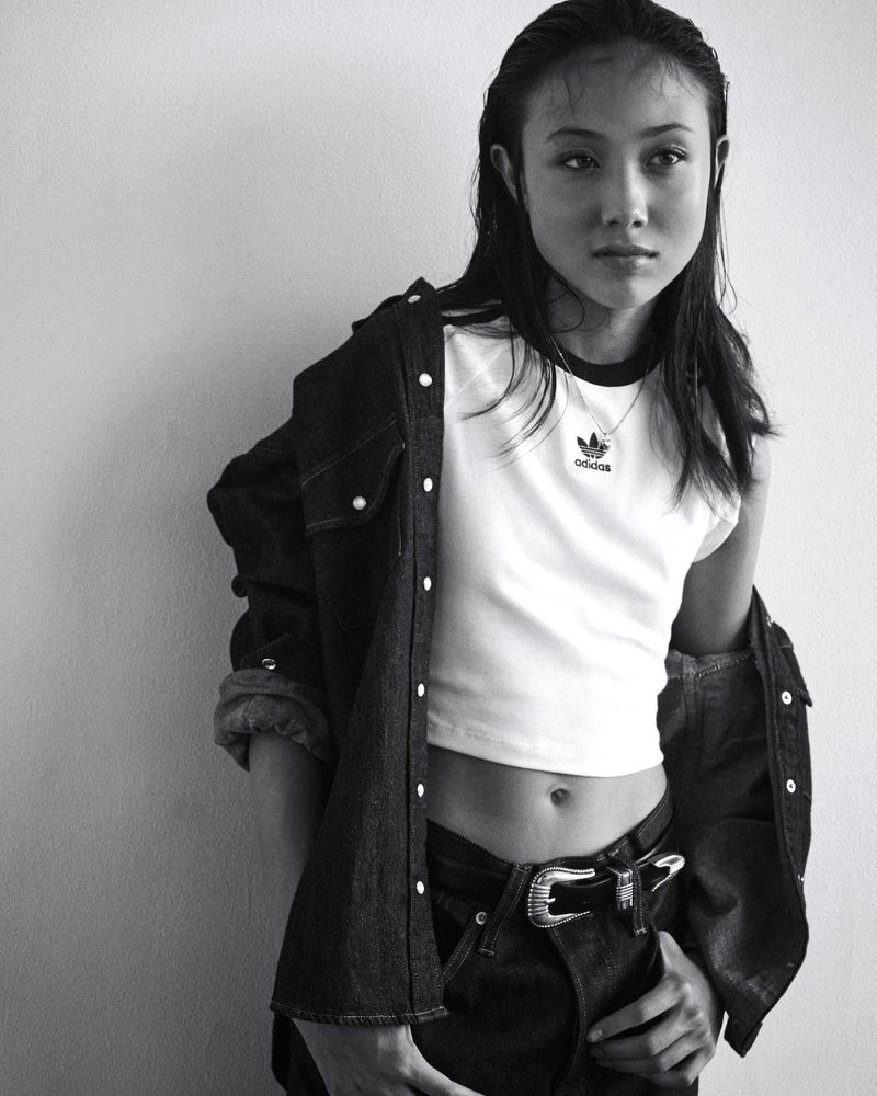 Monochrome portrait of a young adult woman wearing an Adidas t-shirt and denim shirt against a neutral wall indoors.