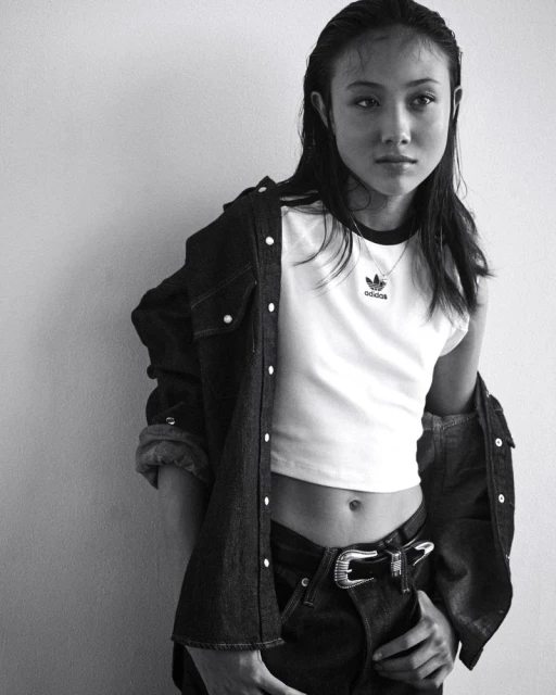Monochrome portrait of a young adult woman wearing an Adidas t-shirt and denim shirt against a neutral wall indoors.