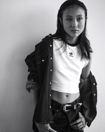 Monochrome portrait of a young adult woman wearing an Adidas t-shirt and denim shirt against a neutral wall indoors.