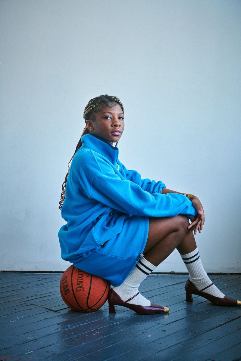 An African American woman in a blue tracksuit sitting on a basketball in a studio, wearing striped socks and heeled Mary Jane shoes.