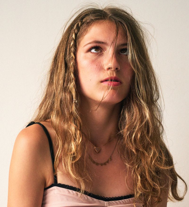 Portrait of a young adult female looking up with a braid in her long wavy hair, wearing a pink top against a white background.