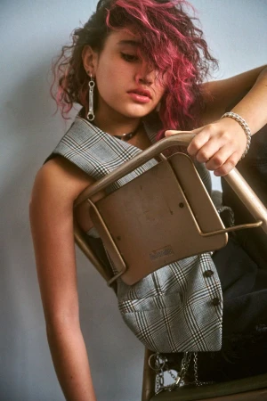 Portrait of a stylish woman with pink-streaked hair, wearing a vest and jewelry, posing indoors with a chair, against a plain wall.