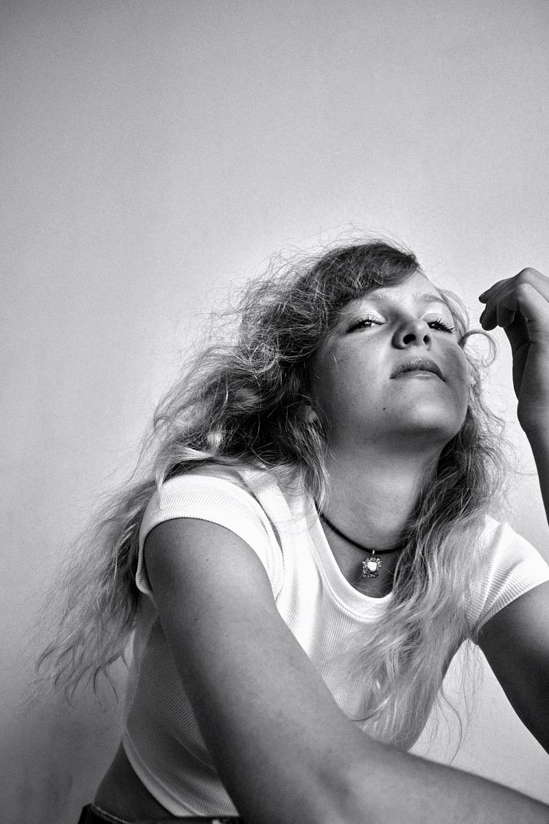 Young woman with curly blonde hair looking upward, wearing a white crop top and pendant necklace, shot in black and white.