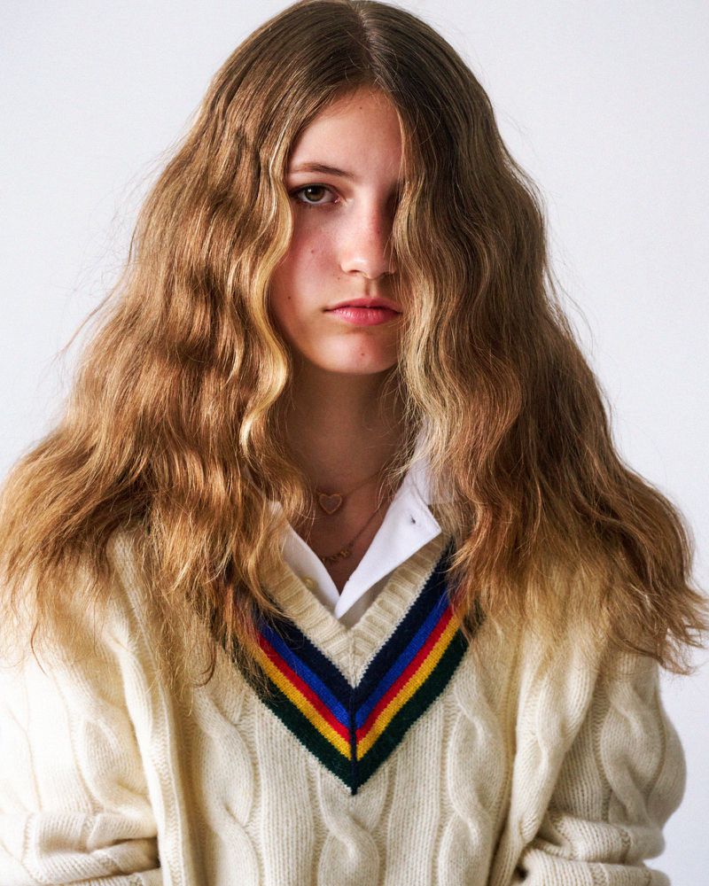 Portrait of a young person with long brown hair, wearing a cream-colored cable knit sweater with a colorful striped V-neck collar against white.