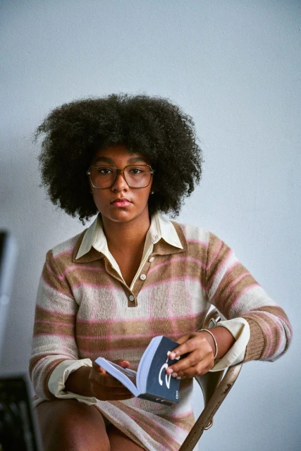 Indoor portrait of a young adult female with an afro, wearing a striped knit outfit and glasses, seated and reading a book.