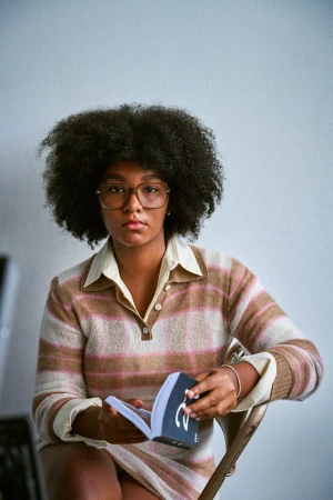 Indoor portrait of a young adult female with an afro, wearing a striped knit outfit and glasses, seated and reading a book.