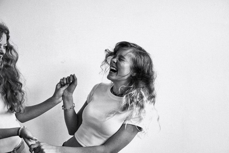 Two women laugh, holding hands in black and white. One woman faces the camera, the other faces away. White background studio shot.