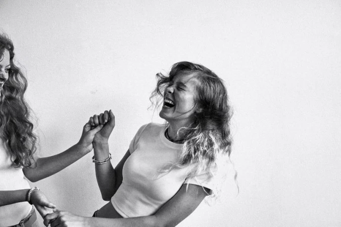 Two women laugh, holding hands in black and white. One woman faces the camera, the other faces away. White background studio shot.