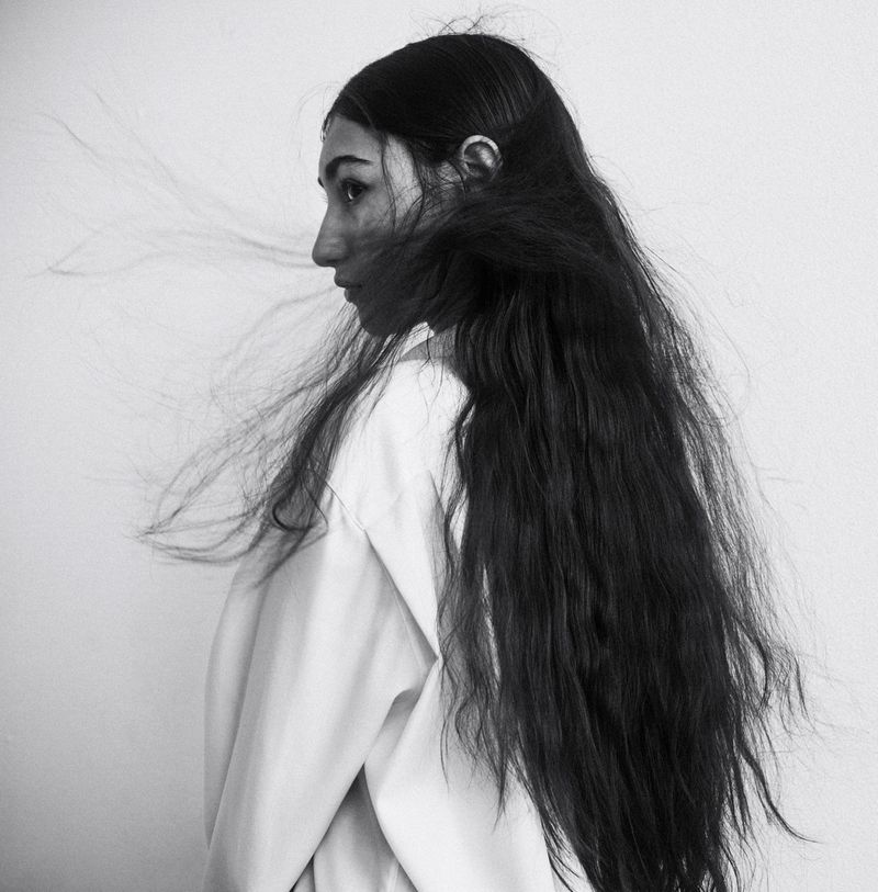Monochrome portrait of a young woman in profile with long, dark hair blowing across her face, wearing a light-colored shirt against a plain background.