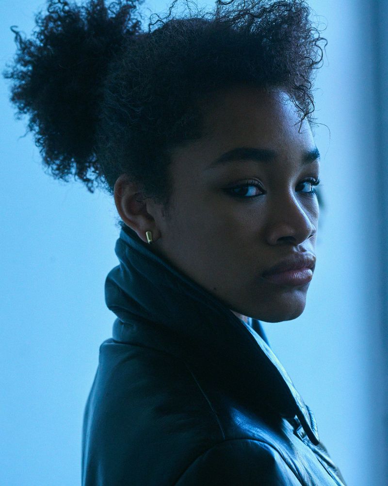 Portrait of a woman with dark hair in a bun wearing a leather jacket, standing against a blue background, looking directly at the camera.