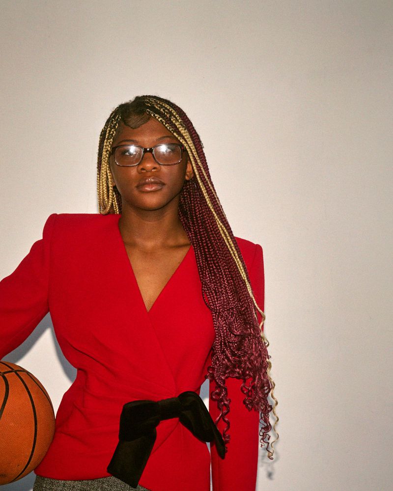 Young woman with long braids in a red blazer holding a basketball poses against a white background in a medium, portrait-oriented shot.