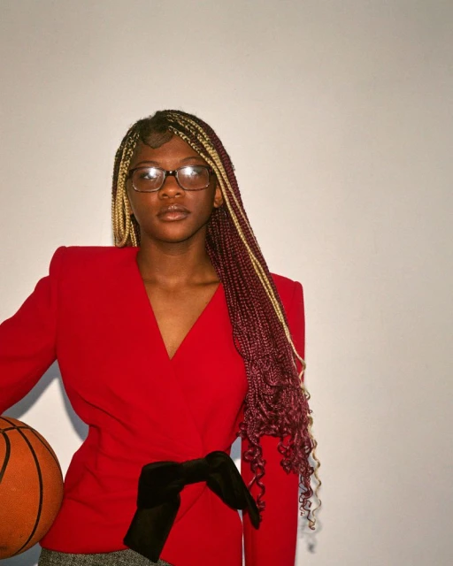 Young woman with long braids in a red blazer holding a basketball poses against a white background in a medium, portrait-oriented shot.