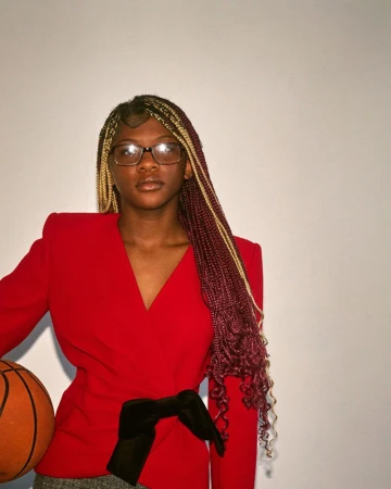 Young woman with long braids in a red blazer holding a basketball poses against a white background in a medium, portrait-oriented shot.