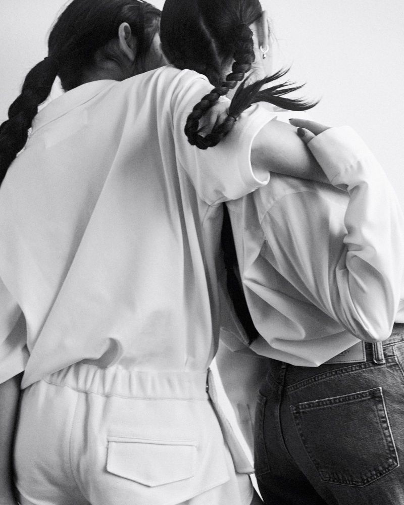 Two women hug indoors in black and white. One has long dark braids and wears a white shirt and pants. The other wears jeans.