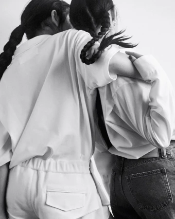 Two women hug indoors in black and white. One has long dark braids and wears a white shirt and pants. The other wears jeans.