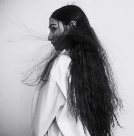 Profile portrait of a young woman with long flowing dark hair blowing in the wind against a plain white background.