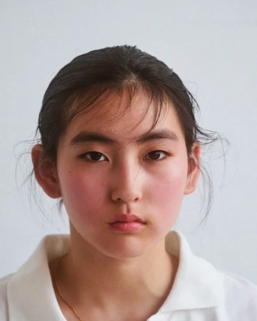 Close-up portrait of a young person with dark hair and rosy cheeks wearing a white collared shirt against a white seamless background