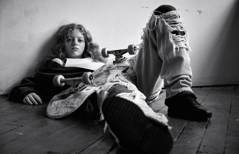 Young person sitting on a wooden floor leaning against a plain wall with a skateboard resting across their legs.