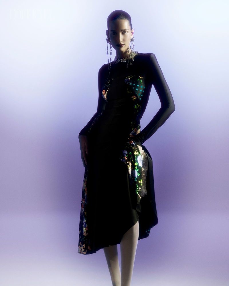 A woman poses in a black dress with colorful patterned accents, accessorized with long earrings and a necklace, against a gradient background.