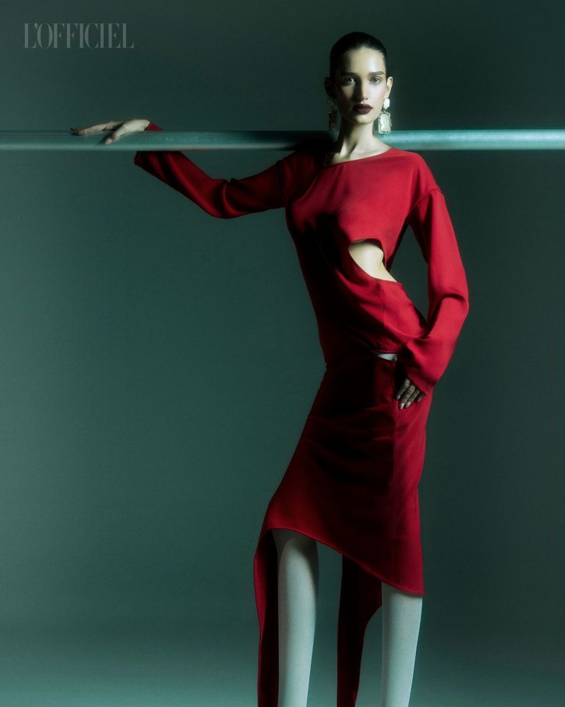 Fashion editorial shot of a slender woman in a red cutout dress with long sleeves, posing near a horizontal metal bar in a studio.