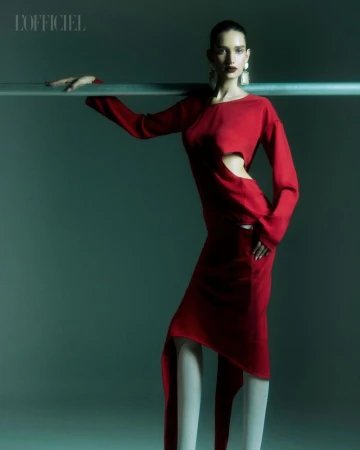 Fashion editorial shot of a slender woman in a red cutout dress with long sleeves, posing near a horizontal metal bar in a studio.