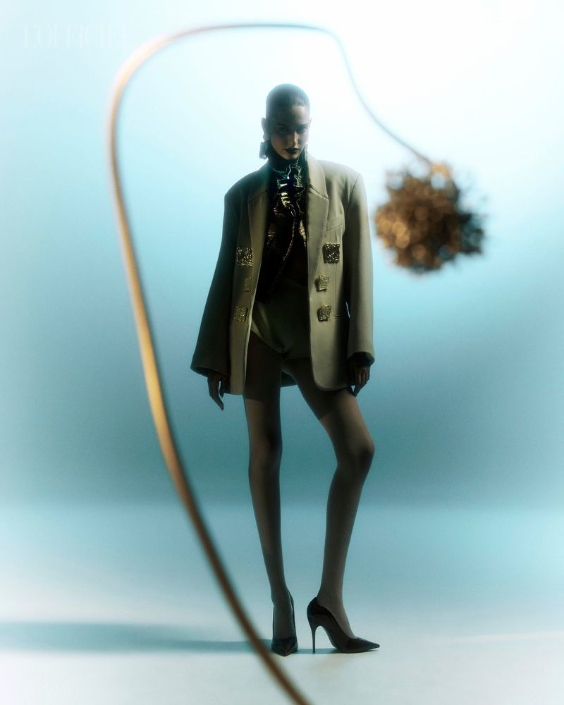A stylish adult female in a blazer, shorts, tights, and heels posing in a photo studio with a decorative grass and neutral-toned gradient background.