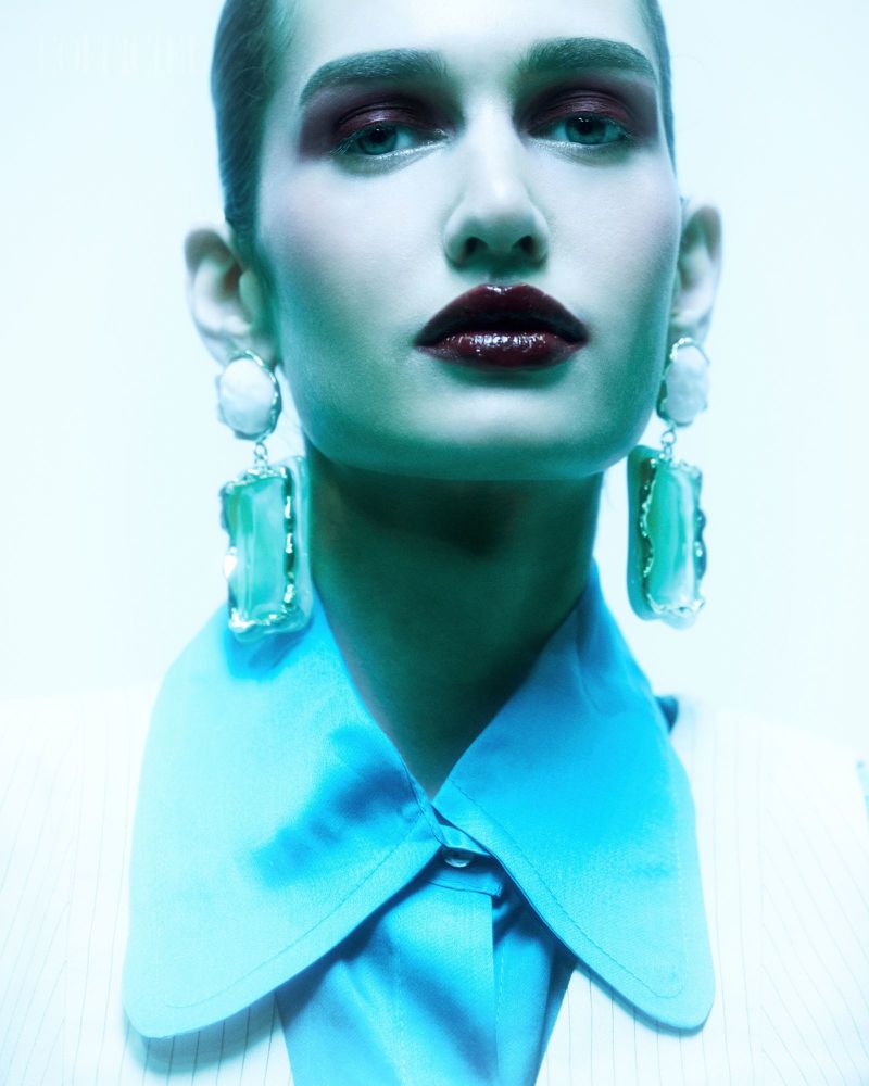Close-up portrait of a woman with dark makeup, prominent earrings, a striking blue collar, and dramatic lighting for a beauty advertisement.