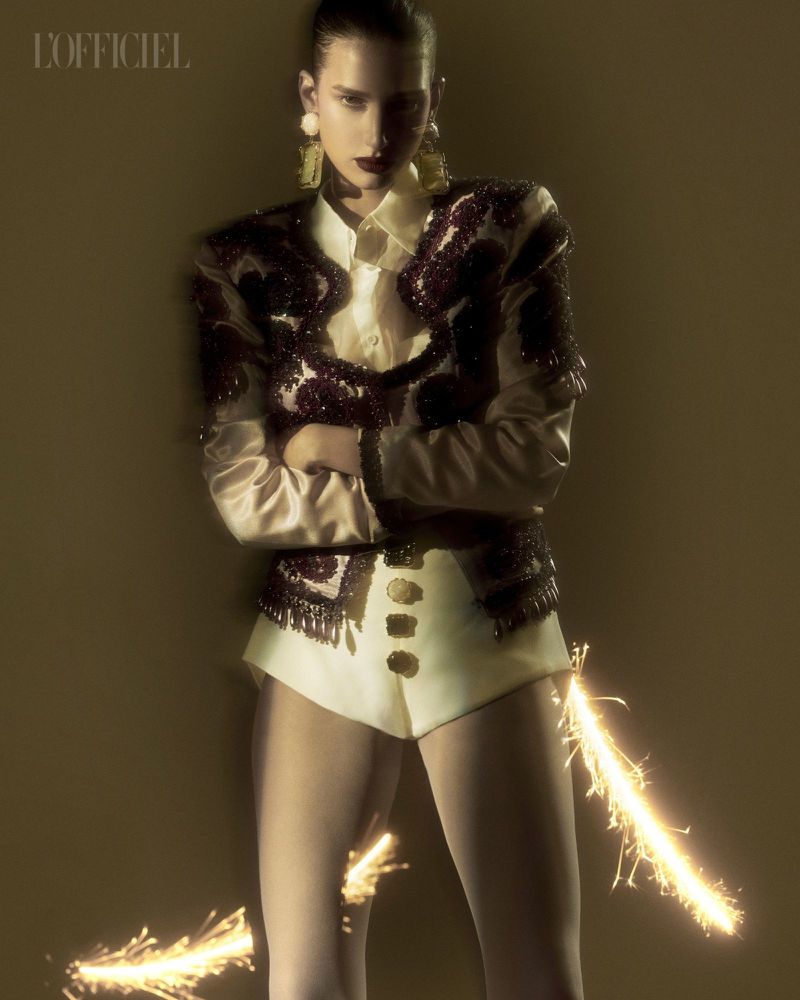 A model in a light-colored outfit poses indoors with crossed arms and decorative earrings. Sparks fly around the lower part of her body.