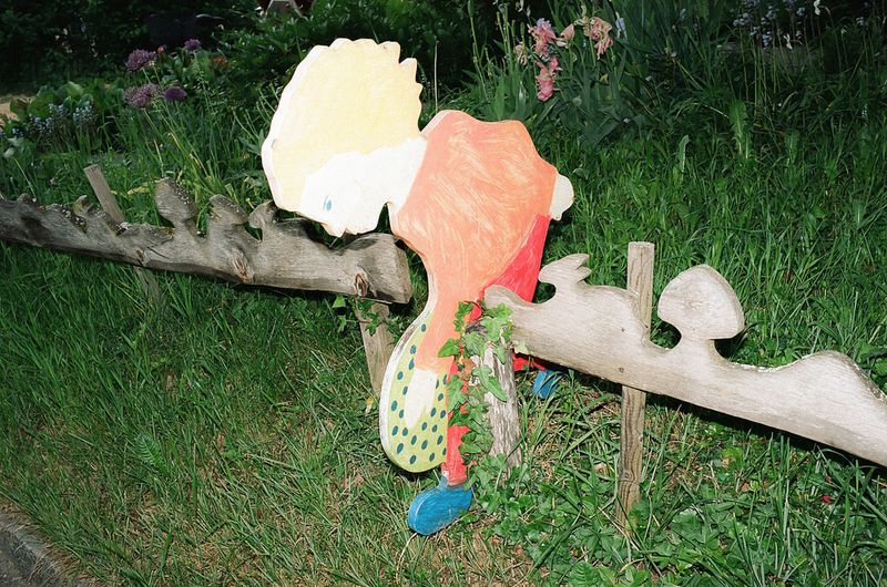 Whimsical garden art: a colorful, painted wooden cutout of a person on a rustic wooden fence surrounded by lush green grass and plants.