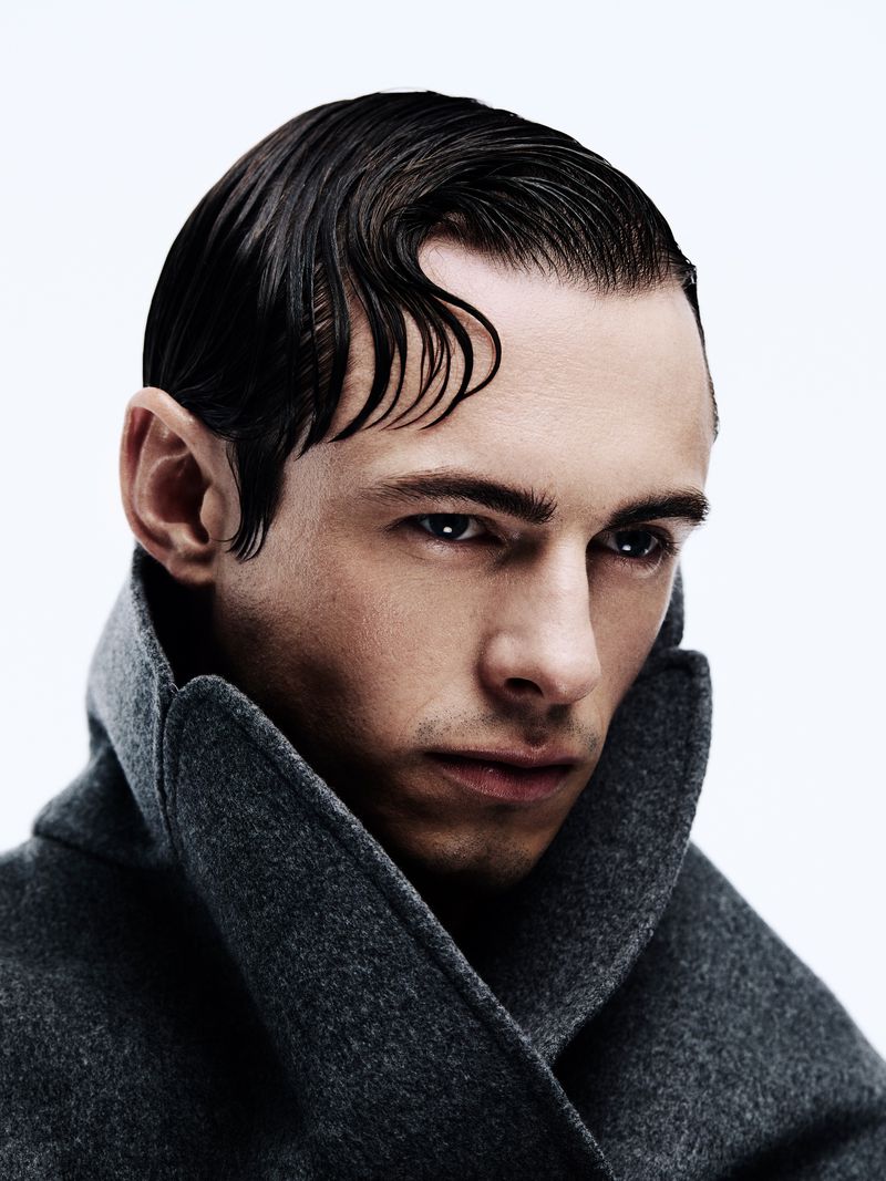 Portrait of a fashionable adult male with slicked-back dark hair and a grey wool coat, posing in a photo studio with bright, even lighting.