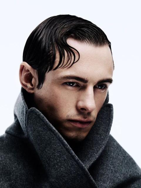 Portrait of a fashionable adult male with slicked-back dark hair and a grey wool coat, posing in a photo studio with bright, even lighting.
