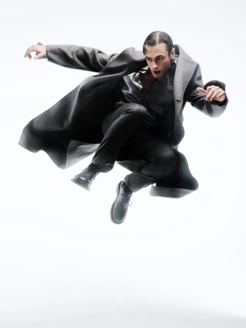 A person in a gray coat mid-air, captured in a dynamic studio shot on a white backdrop, showcasing fashion and movement in a single frame.