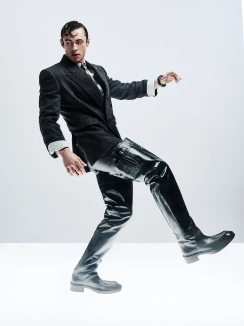 Fashion photo of a man in a suit and tall black boots posing in a studio. Dramatic lighting highlights textures of fabric and leather.