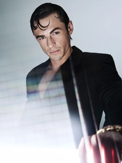 Stylish man in dark jacket poses in a studio with bright lighting. He looks at the camera with slicked-back hair and a bracelet.